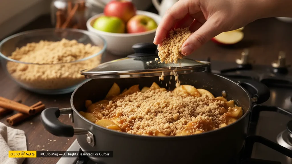 پاشیدن مخلوط کرامبل روی سیبهای پخته شده در یک قابلمه روی اجاق گاز.